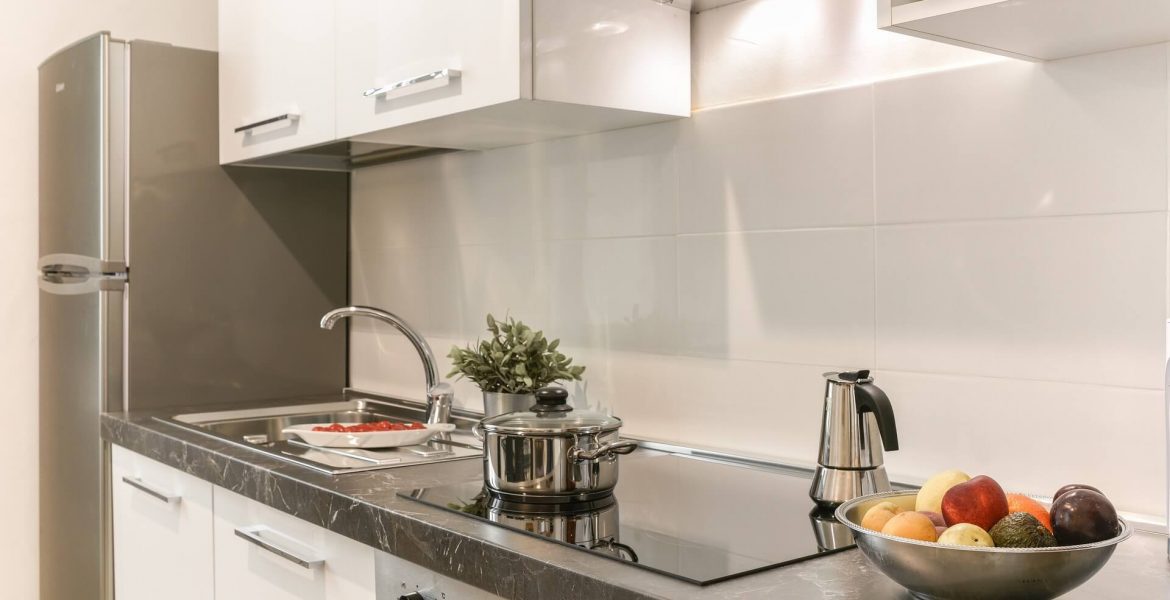 Białe szafki ze srebrnymi uchwytami w kuchni, srebrny garnek stoi na płycie indukcyjnej, obok srebrna kawiarka, owoce i zlew.