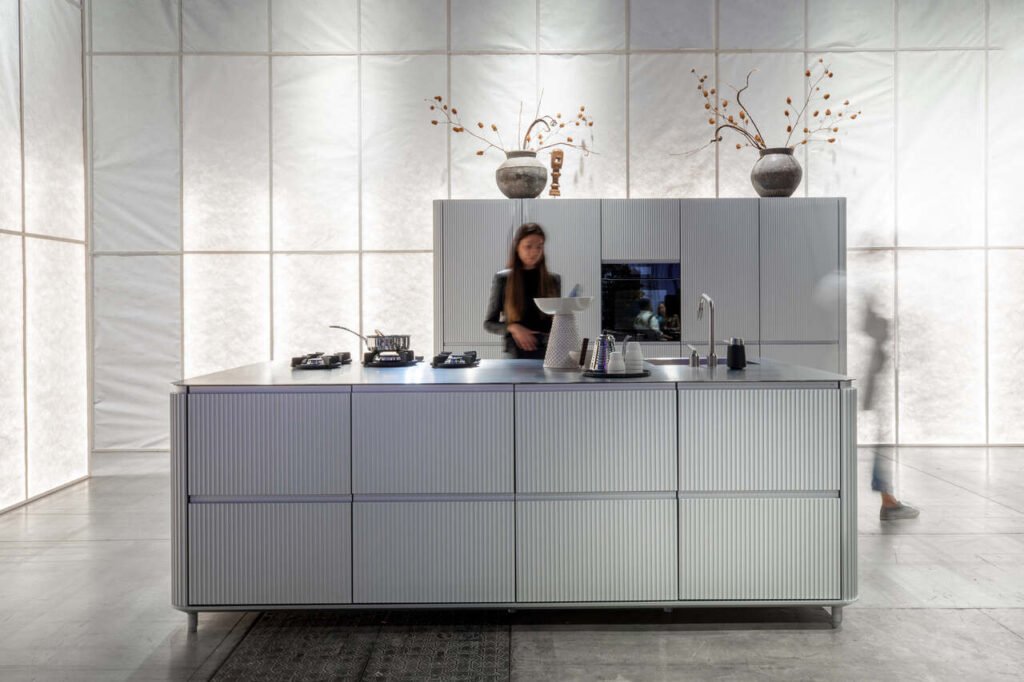 V3 jest hołdem dla 85. rocznicy powstania Vipp , duńskiej marki specjalizującej się w obróbce metali. Samonośne moduły są pokryte naturalnym anodyzowanym aluminium, z pionowo wytłaczanymi profilami, które zakrzywiają się wzdłuż krawędzi, nadając wykończeniu z efektem metalu subtelną elegancję.  Fot. Monica Spezia | Salone del Mobile.Milano.