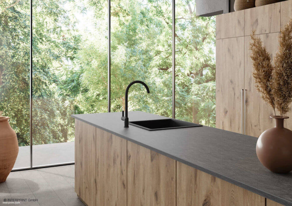 Szary dekor Xisto firmy Interprint użyty na blacie w kuchni, kuchnia w kolorze jasnego drewna, czarny zlew i bateria kuchenna, w tle zielone drzewa - miesięcznik i portal informacyjny branży meblarskiej biznes.meble.pl