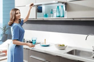 Amix - Pani w niebieskiej sukience otwierająca szafkę górną z systemem Top-Stays SQ w szarej kuchni - miesięcznik i portal informacyjny branży meblarskiej biznes.meble.pl