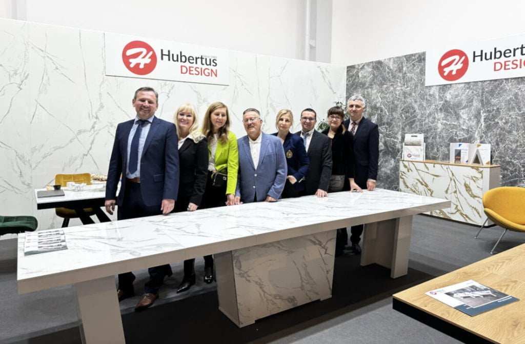 Cały zespół firmy Hubertus Design, osiem osób stojących przy dużym stole, na stoisku podczas targów Meble Polska 2023 w Poznaniu - miesięcznik i portal informacyjny branży meblarskiej