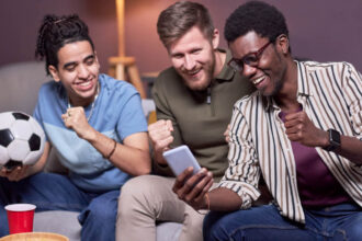 Orion Media - bukmacher, zakłady, zawód Typer - trzech mężczyzn siedzących na kanapie oglądających mecz na żywo przez smartfon