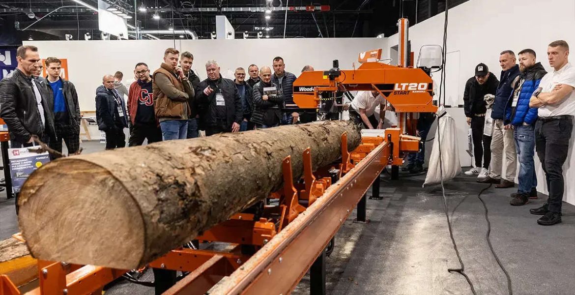 Grupa osób obserwujących pokaz przecinania pnia drzewa na profesjonalnej traku taśmowym podczas targów technologii obróbki drewna.
