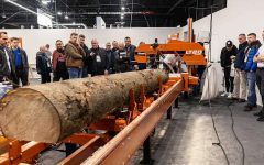 Grupa osób obserwujących pokaz przecinania pnia drzewa na profesjonalnej traku taśmowym podczas targów technologii obróbki drewna.