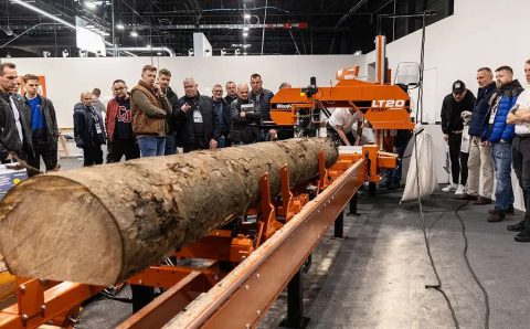 Grupa osób obserwujących pokaz przecinania pnia drzewa na profesjonalnej traku taśmowym podczas targów technologii obróbki drewna.