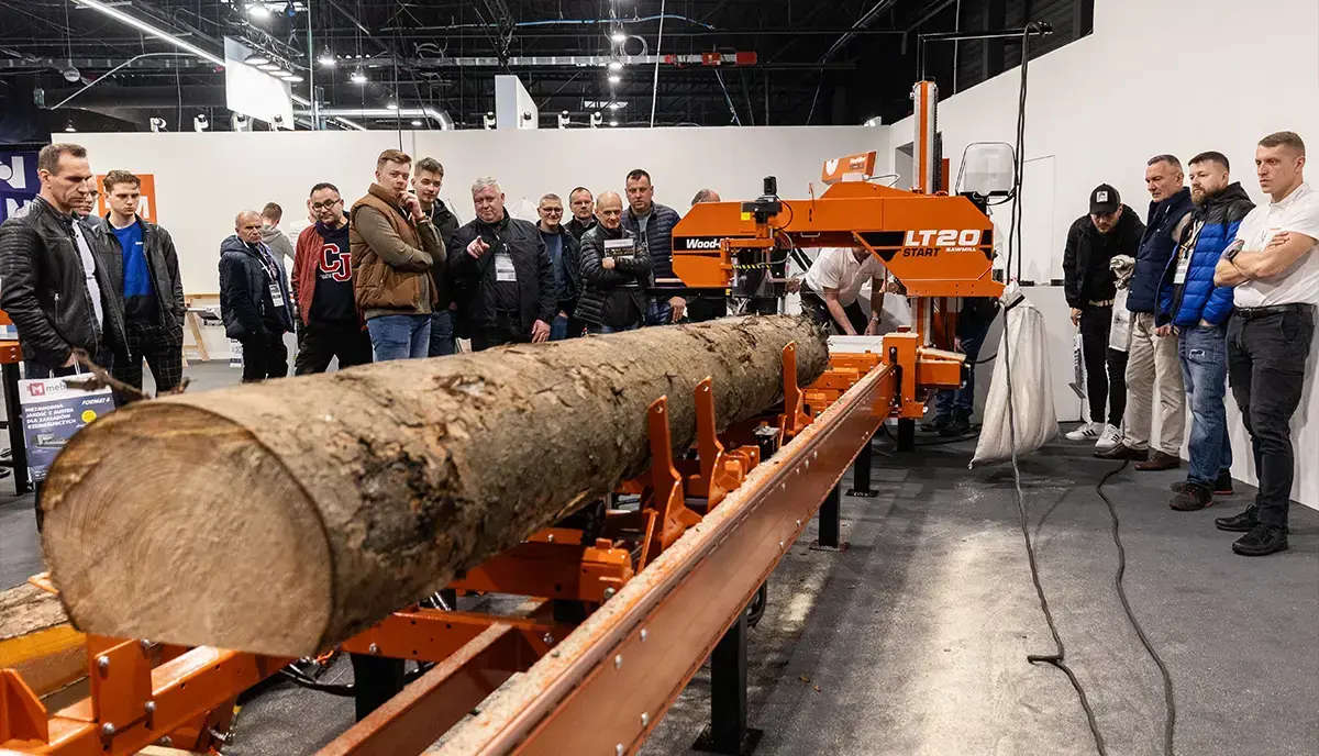 Grupa osób obserwujących pokaz przecinania pnia drzewa na profesjonalnej traku taśmowym podczas targów technologii obróbki drewna.