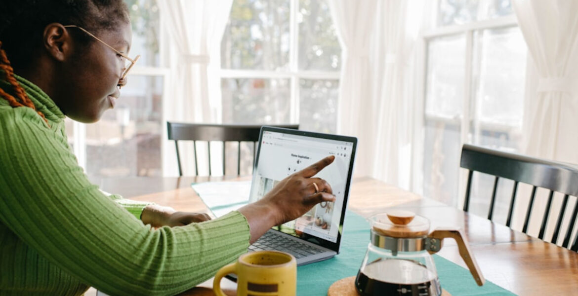 Ubezpieczenie mieszkania - kobieta siedzi przy stoliku z kubkiem kawy i wskazuje na ekran laptopa.