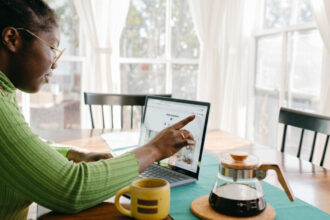 Ubezpieczenie mieszkania - kobieta siedzi przy stoliku z kubkiem kawy i wskazuje na ekran laptopa.
