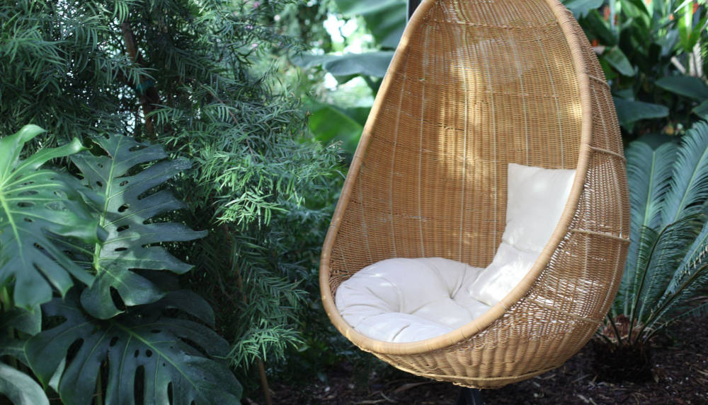 Pleciony, wiszący kokon. fotel ogrodowy wśród ciemnozielonej roślinności.