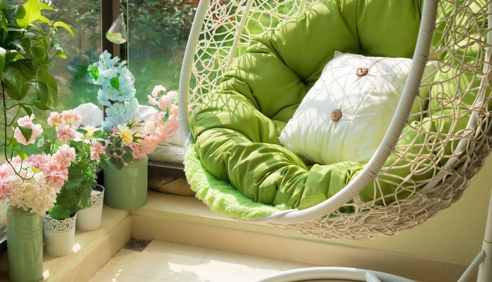 Pleciony wiszący kokon, fotel ogrodowy w jasnej kolorystyce z zielonymi poduszkami na słonecznym tarasie.