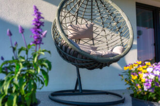 Pleciony wiszący kokon, fotel ogrodowy na tarasie wśród kwiatów.