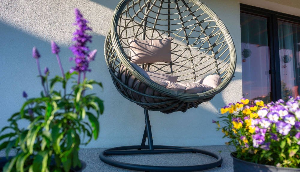 Pleciony wiszący kokon, fotel ogrodowy na tarasie wśród kwiatów.