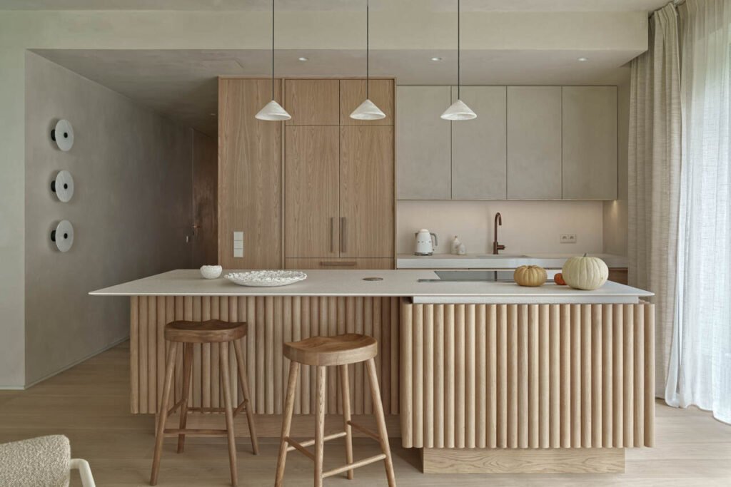 Apartament wakacyjny nad polskim morzem o powierzchni 230 m2. Kuchnia wykonana z forniru dębowego z blatami z materiału lapitec o dużej strukturze. Szafki wiszące obłożone tynkiem dekoracyjnym, tak samo jak ściany i sufity. Lampy nad wyspą i kinkiety na ścianie z alabastru. Hokery przy wyspie – Zara Home.