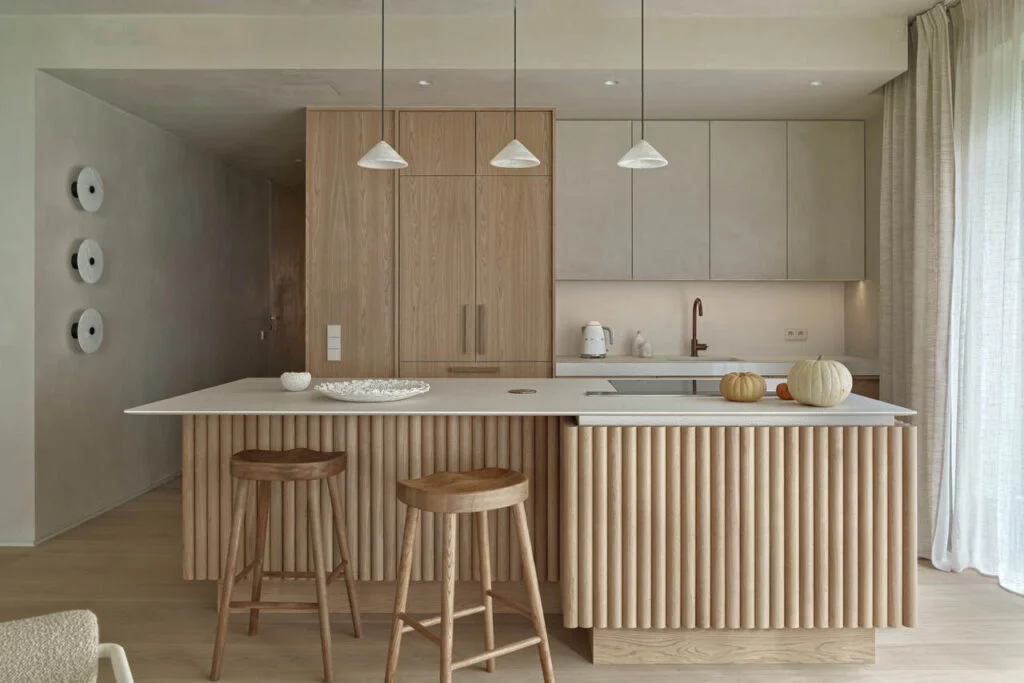 Apartament wakacyjny nad polskim morzem o powierzchni 230 m2. Kuchnia wykonana z forniru dębowego z blatami z materiału lapitec o dużej strukturze. Szafki wiszące obłożone tynkiem dekoracyjnym, tak samo jak ściany i sufity. Lampy nad wyspą i kinkiety na ścianie z alabastru. Hokery przy wyspie – Zara Home.