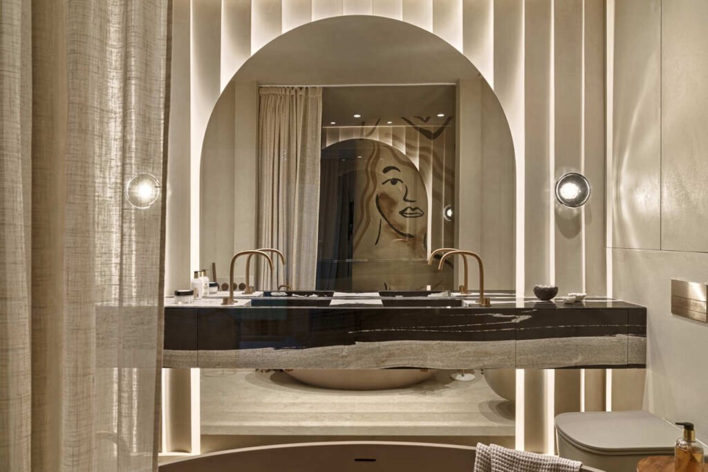 Wnętrze łazienki w apartamencie wakacyjnym. Blat kamienny z granitu copacabana, baterie miedziane firmy Cocoon, lustro z podświetleniem na tle ściany wykonanej z giętej płyty karton gipsowej i obłożone tynkiem dekoracyjnym Kerrakol Design. W kamiennym blacie po lewej i prawej stronie znajdują się szuflady na kosmetyki, suszarkę itp.