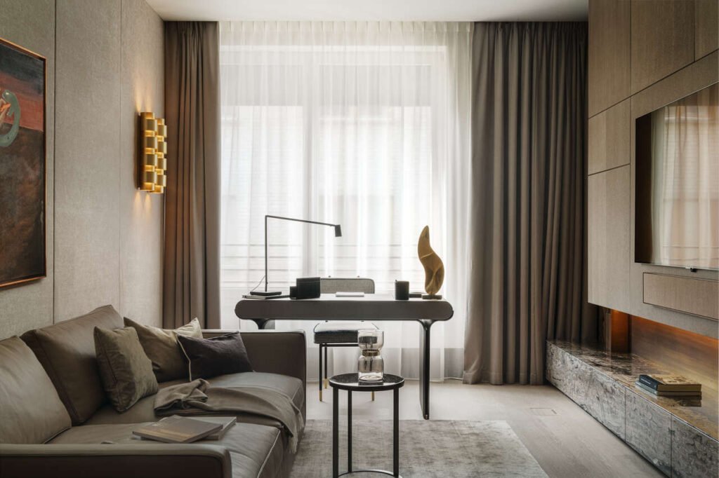 Salonik przy sypialni w apartamencie w Warszawie. Biurko Galootti & Radice, sofa B&B Italia z brązowej skóry. Zabudowa po prawej z telewizorem z forniru marki Tabu, blaszaną okładziną nad półka i półką z szufladami obłożoną kamieniem cafe late. Po lewej na ścianie tapicerka i kinkiety marki CTO.