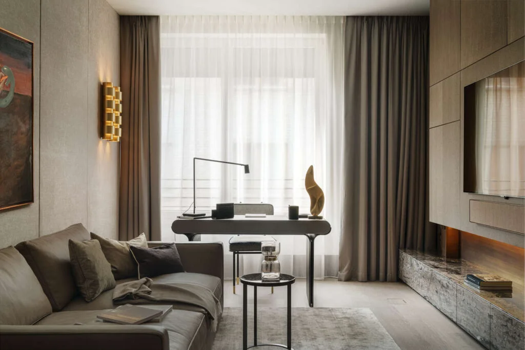 Salonik przy sypialni w apartamencie w Warszawie. Biurko Galootti & Radice, sofa B&B Italia z brązowej skóry. Zabudowa po prawej z telewizorem z forniru marki Tabu, blaszaną okładziną nad półka i półką z szufladami obłożoną kamieniem cafe late. Po lewej na ścianie tapicerka i kinkiety marki CTO.