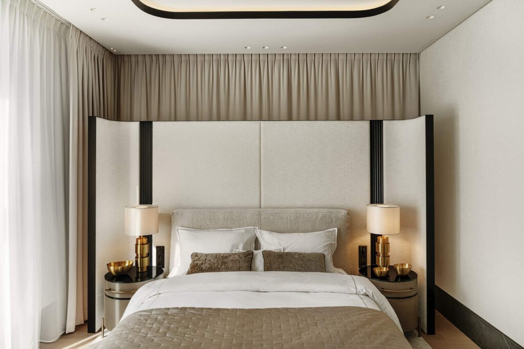 Sypialnia. Za łóżkiem znajduje się okno zasłonięte zasłoną, a na jego tle parawan tapicerowany z łóżkiem marki Flou, stolikami nocnymi marki Baxter, lampkami marki CTO oraz tapetą na ścianach marki Casamance. We wnętrzu wyróżniają się czarne dodatki przy tapicerce oraz w suficie.