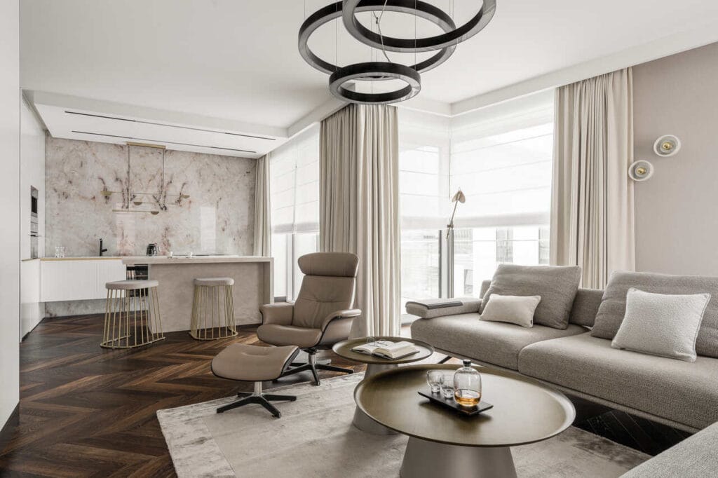 Strefa wypoczynkowa z aneksem kuchennym w apartamencie wakacyjnym w inwestycji Yacht Park. Gdy łączymy elementy modern classic z nowoczesnością powstaje eleganckie, ponadczasowe wnętrze, bez zbędnego przeładowania i dekoracji, ale w wysokiej klasy materiałach. Połączenie naturalnego onyxu, który od tyłu ma dodatkowo podświetlenie ledowe, z pięknymi beżami spieków kwarcowych Laminam Cava Diamond Cream. Ciemna, wędzona podłoga Kaczkan, sofa Olta, stoliki Cattelan Italia, fotel Conform, lampy w strefie dziennej Olev, lampa w kuchni Bolia. Fot. Katarzyna Seliga-Wróblewska, Marcin Wróblewski / Fotomohito.