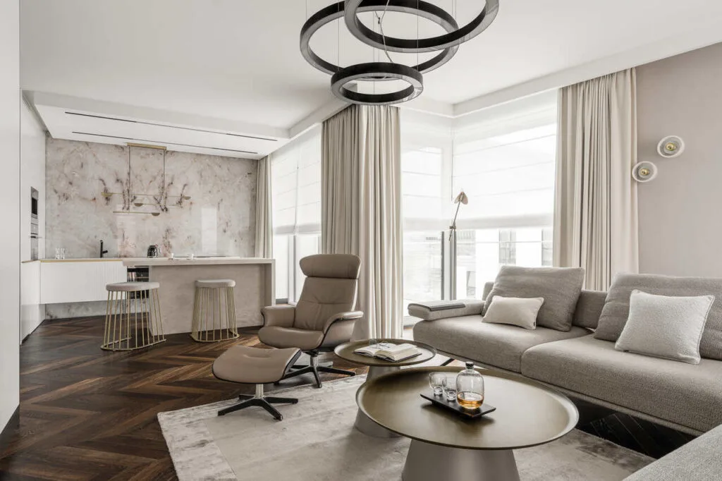 Strefa wypoczynkowa z aneksem kuchennym w apartamencie wakacyjnym w inwestycji Yacht Park. Gdy łączymy elementy modern classic z nowoczesnością powstaje eleganckie, ponadczasowe wnętrze, bez zbędnego przeładowania i dekoracji, ale w wysokiej klasy materiałach. Połączenie naturalnego onyxu, który od tyłu ma dodatkowo podświetlenie ledowe, z pięknymi beżami spieków kwarcowych Laminam Cava Diamond Cream. Ciemna, wędzona podłoga Kaczkan, sofa Olta, stoliki Cattelan Italia, fotel Conform, lampy w strefie dziennej Olev, lampa w kuchni Bolia. Fot. Katarzyna Seliga-Wróblewska, Marcin Wróblewski / Fotomohito.