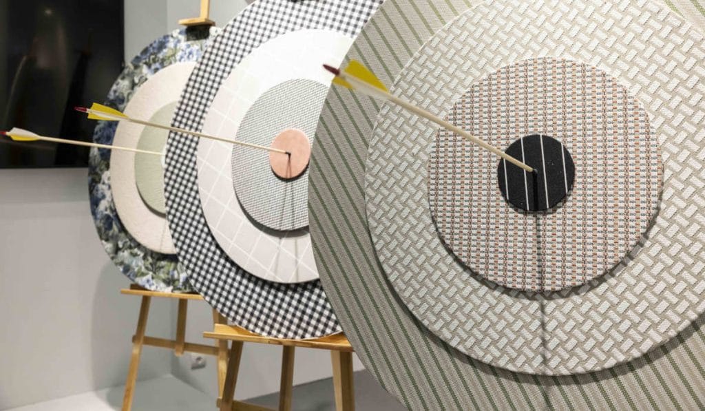 Trzy tarcze strzelnicze wykonane z różnych tkanin z wbitymi strzałami, prezentacja wzorów materiałów na stojakach podczas targów tekstylnych.