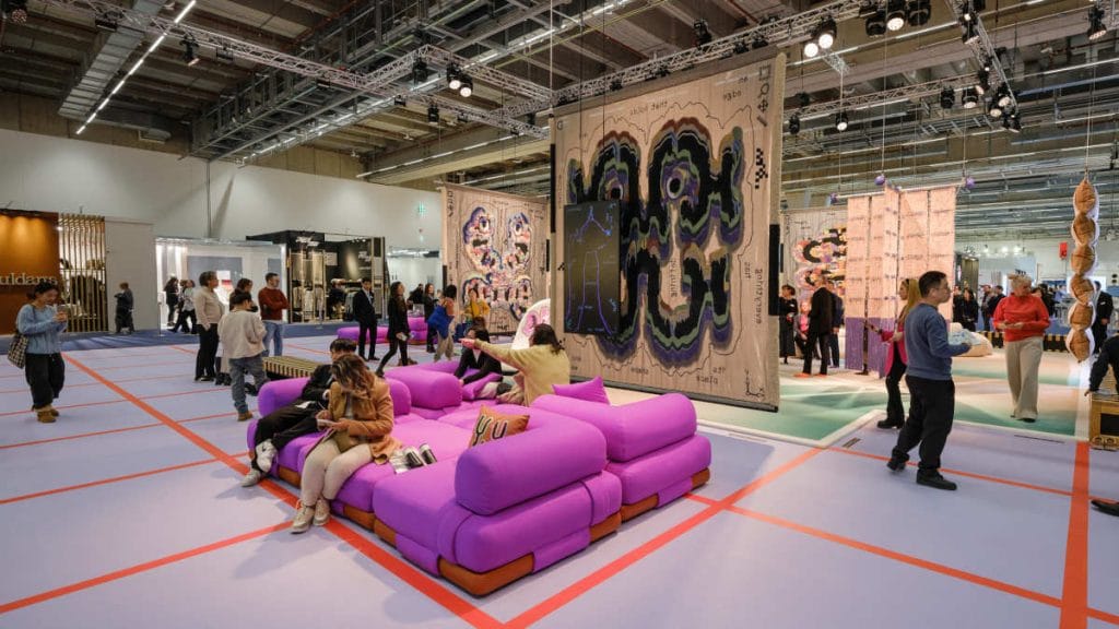 Zwiedzający spacerujący po kolorowej hali targowej z fioletowymi sofami i dużymi, artystycznymi tkaninami wiszącymi na ścianach podczas targów wnętrzarskich.