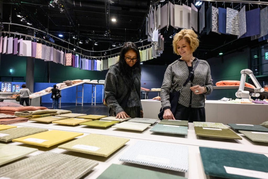 Dwie kobiety oglądające próbki tkanin w różnych kolorach na stole ekspozycyjnym podczas targów tekstylnych, w tle zawieszone próbki materiałów.
