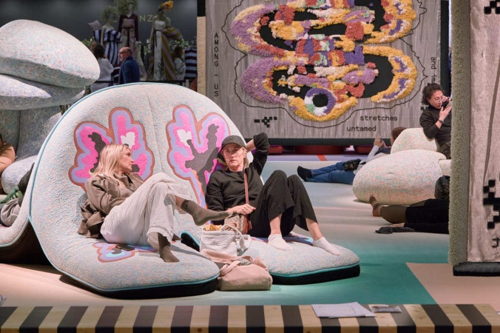 Dwie osoby siedzące na  szaro-fioletowych sofach. W tle artystyczne tkaniny wiszące na ścianach podczas targów wnętrzarskich.