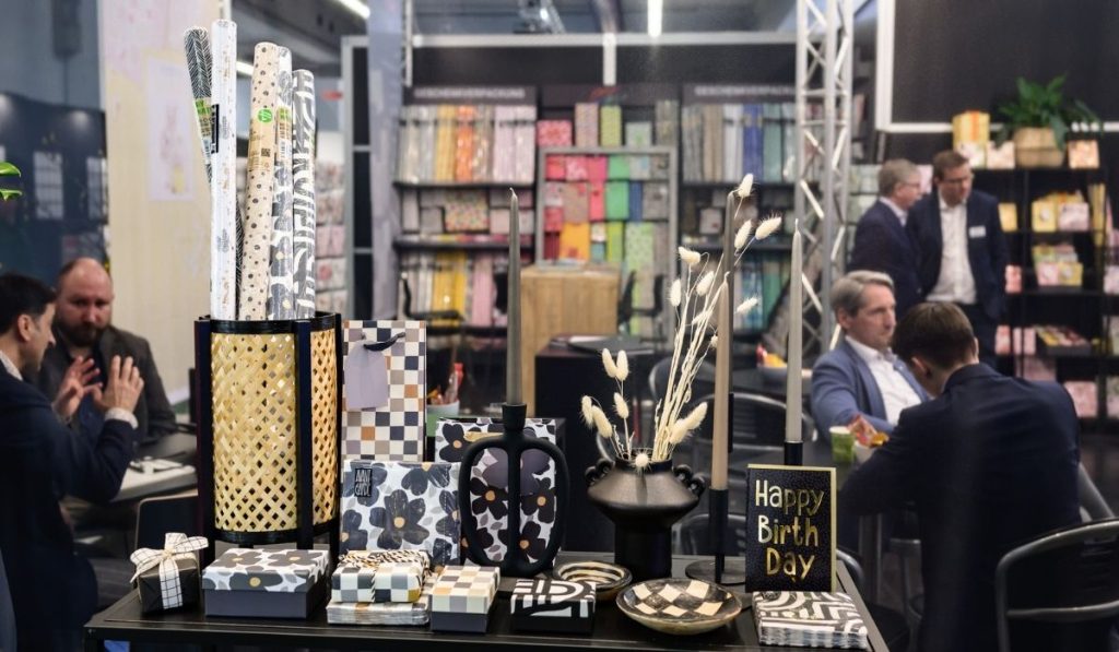 Stoisko targowe z produktami prezentowanymi na pierwszym planie, w tym ozdobnymi papierami do pakowania prezentów w bambusowym koszu, eleganckimi wazonami w czarno-białe i kwiatowe wzory, oraz zestawem prezentowym. W tle widoczne regały z kolorowymi papierami i ludzie prowadzący rozmowy biznesowe.