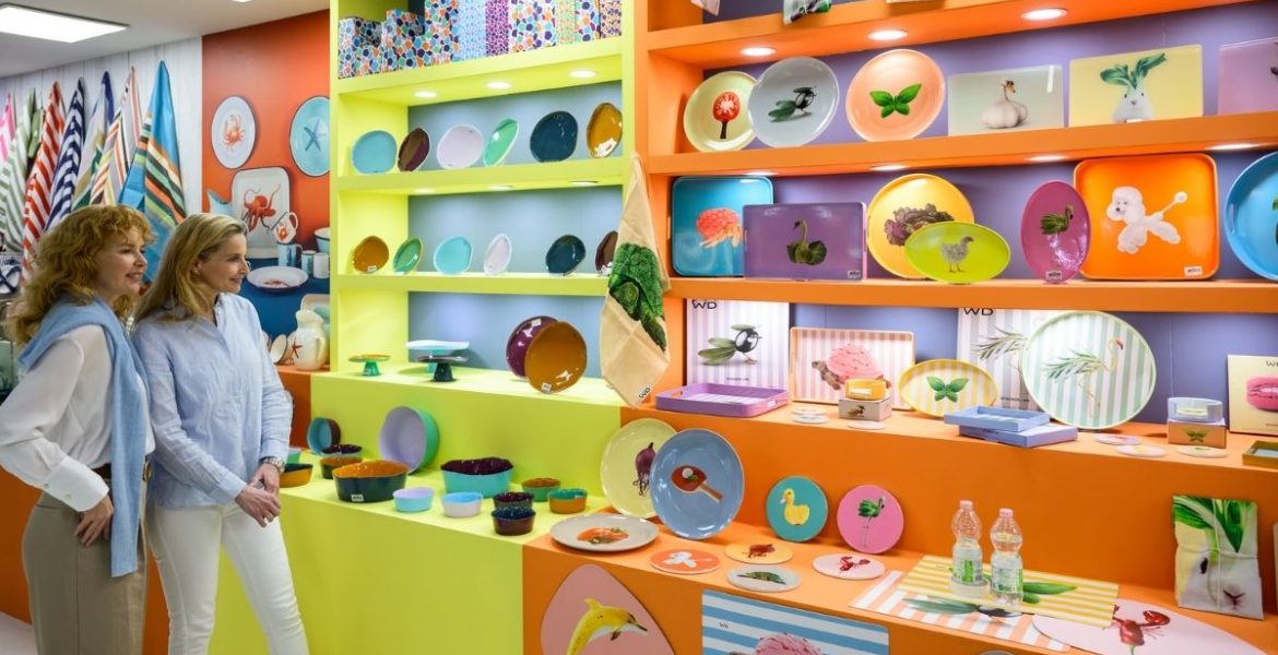 Dwóch kobiet oglądających kolorowe naczynia ceramiczne i talerze dekorowane w jaskrawych, popartowych barwach, przedstawiające motywy zwierzęce i roślinne. Naczynia są wyeksponowane na półkach w kolorach neonowej żółci i pomarańczy.