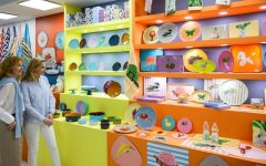 Dwóch kobiet oglądających kolorowe naczynia ceramiczne i talerze dekorowane w jaskrawych, popartowych barwach, przedstawiające motywy zwierzęce i roślinne. Naczynia są wyeksponowane na półkach w kolorach neonowej żółci i pomarańczy.