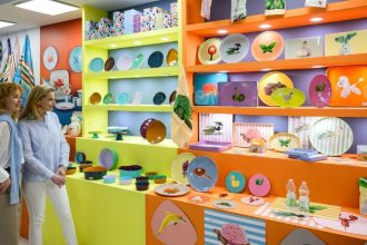 Dwóch kobiet oglądających kolorowe naczynia ceramiczne i talerze dekorowane w jaskrawych, popartowych barwach, przedstawiające motywy zwierzęce i roślinne. Naczynia są wyeksponowane na półkach w kolorach neonowej żółci i pomarańczy.