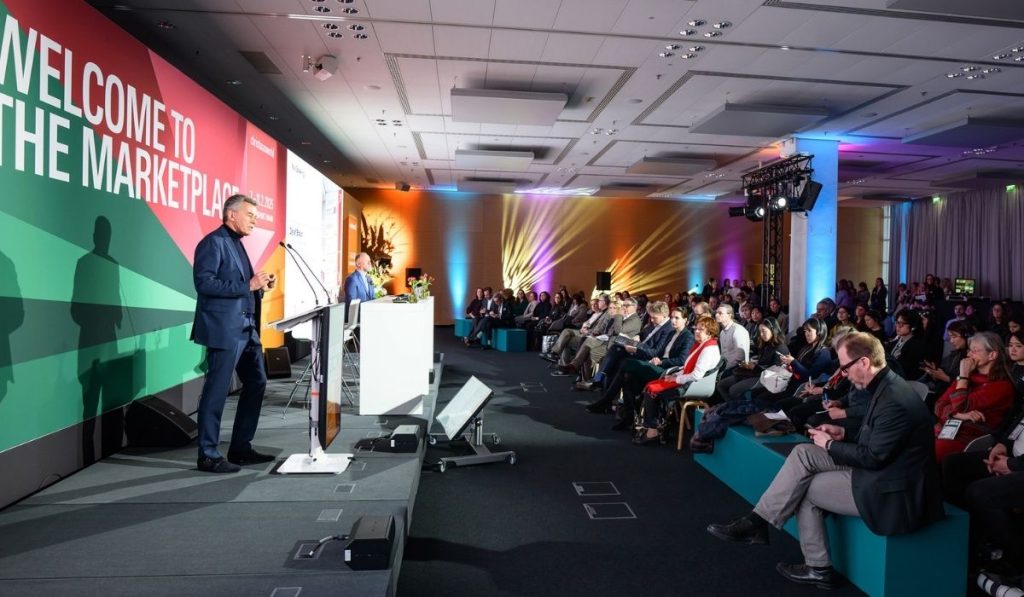 Konferencja prasowa lub wykład z prelegentem w niebieskim garniturze stojącym na scenie przy mównicy. Za nim znajduje się duży ekran z napisem "WELCOME TO THE MARKETPLACE". Na widowni siedzi duża grupa słuchaczy.