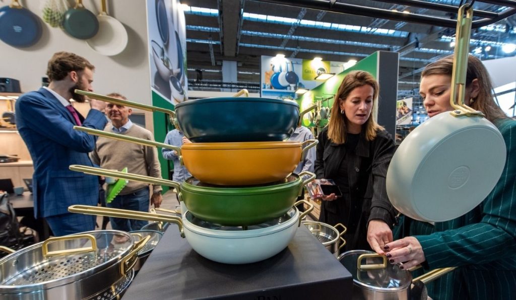 Dwie kobiety oglądające stos kolorowych patelni i rondli z jasnozłotymi uchwytami, eksponowanych na stoisku z wyposażeniem kuchennym. Naczynia są w kolorach morskiego błękitu, musztardowej żółci i butelkowej zieleni.