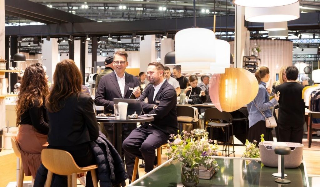 Grupa osób, w tym dwóch mężczyzn w garniturach, siedzących przy okrągłym stole i rozmawiających w otoczeniu nowoczesnego stoiska targowego. Nad nimi wiszą dekoracyjne lampy z dużymi, białymi i drewnianymi abażurami, oświetlające przestrzeń.