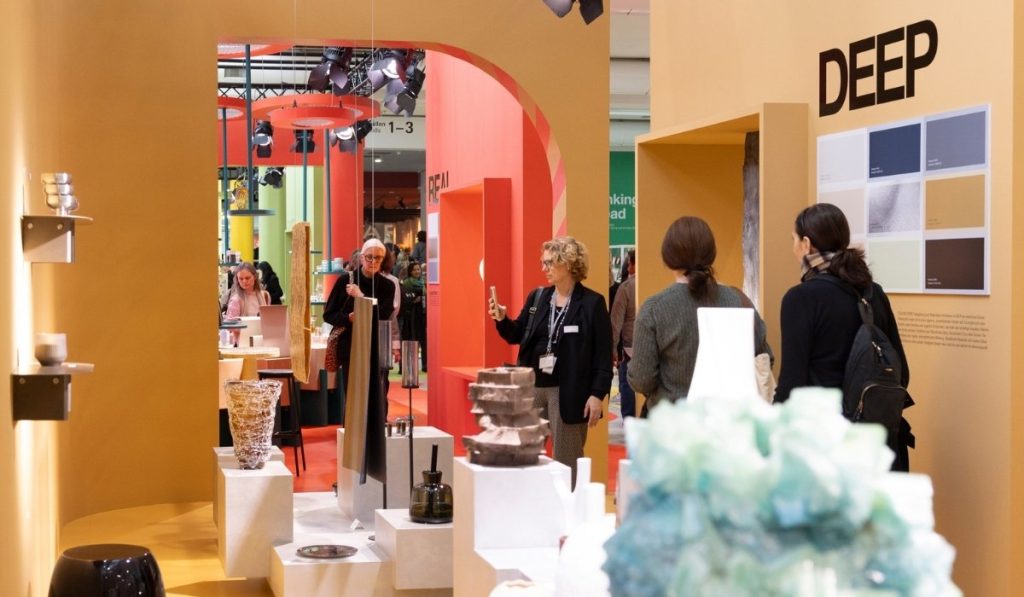 Osoby w strefie trendów na targach, przechodzące przez łukowate, musztardowe przejście. Na pierwszym planie na postumentach eksponowane są nowoczesne, szklane naczynia, a na ścianie widoczny jest napis "DEEP" i paleta kolorów z inspiracjami do wnętrz.