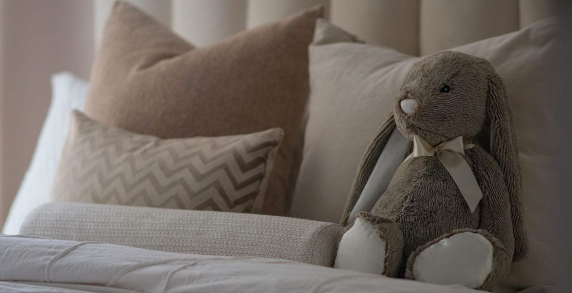 Pluszowy króliczek z oferty Decority siedzi na poduszkach. Zdjęcie w kolorystyce beżowej.