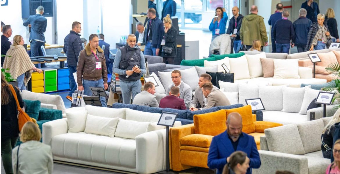 Zabiegany parkiet wystawowy z szeroką ekspozycją nowoczesnych sof i wypoczynków. Uczestnicy targów (kupcy, producenci, projektanci) oglądają i dyskutują o prezentowanych meblach, m.in. na białym i musztardowym narożniku.