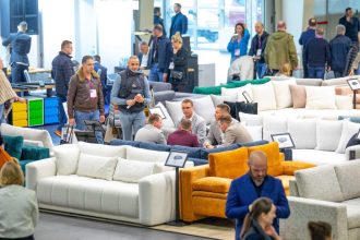 Zabiegany parkiet wystawowy z szeroką ekspozycją nowoczesnych sof i wypoczynków. Uczestnicy targów (kupcy, producenci, projektanci) oglądają i dyskutują o prezentowanych meblach, m.in. na białym i musztardowym narożniku.