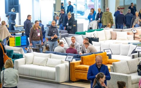 Zabiegany parkiet wystawowy z szeroką ekspozycją nowoczesnych sof i wypoczynków. Uczestnicy targów (kupcy, producenci, projektanci) oglądają i dyskutują o prezentowanych meblach, m.in. na białym i musztardowym narożniku.