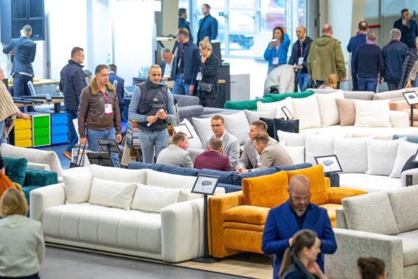 Zabiegany parkiet wystawowy z szeroką ekspozycją nowoczesnych sof i wypoczynków. Uczestnicy targów (kupcy, producenci, projektanci) oglądają i dyskutują o prezentowanych meblach, m.in. na białym i musztardowym narożniku.