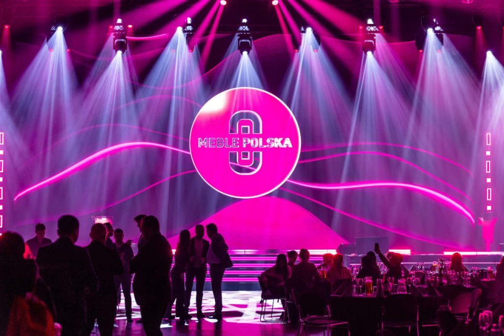 Dynamiczna sceneria bankietu lub gali podczas targów Meble Polska, z dominującym neonowo-różowym logo na tle sceny oświetlonej reflektorami, tworząca uroczystą i nowoczesną atmosferę.