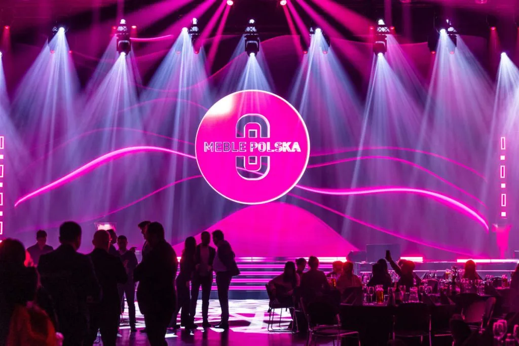 Dynamiczna sceneria bankietu lub gali podczas targów Meble Polska, z dominującym neonowo-różowym logo na tle sceny oświetlonej reflektorami, tworząca uroczystą i nowoczesną atmosferę.