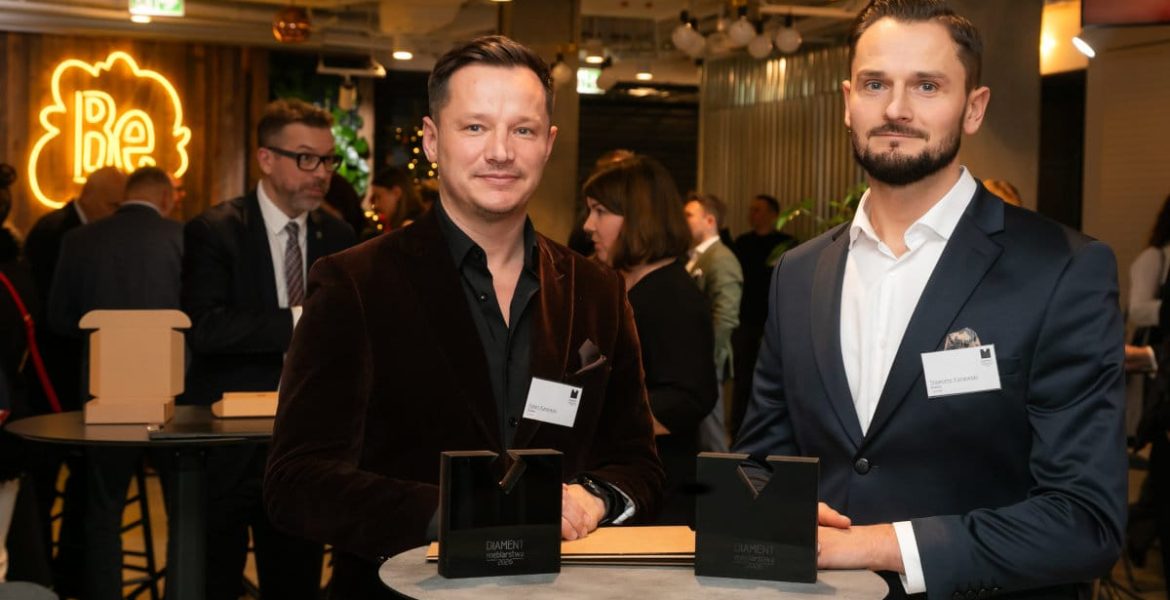 Dwaj mężczyźni w eleganckich marynarkach stoją przy okrągłym stoliku podczas gali meblarskiej. Przed nimi na blacie leżą dwie czarne statuetki „Diament Meblarstwa 2026”. W tle widoczne są inne osoby oraz neonowy logotyp.