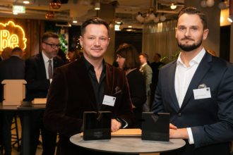 Dwaj mężczyźni w eleganckich marynarkach stoją przy okrągłym stoliku podczas gali meblarskiej. Przed nimi na blacie leżą dwie czarne statuetki „Diament Meblarstwa 2026”. W tle widoczne są inne osoby oraz neonowy logotyp.