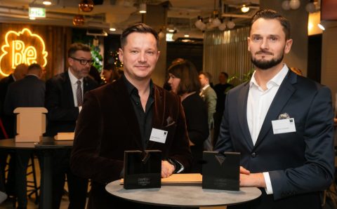 Dwaj mężczyźni w eleganckich marynarkach stoją przy okrągłym stoliku podczas gali meblarskiej. Przed nimi na blacie leżą dwie czarne statuetki „Diament Meblarstwa 2026”. W tle widoczne są inne osoby oraz neonowy logotyp.