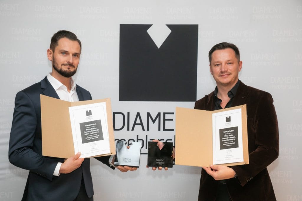 Dwaj mężczyźni prezentują otrzymane nagrody na tle ścianki z logo „DIAMENT meblarstwa”. Każdy z nich trzyma w dłoniach otwarty dyplom w teczce oraz czarną statuetkę. Napisy na dyplomach wskazują na nagrodę dla Kolekcji Modulo firmy Brattex.