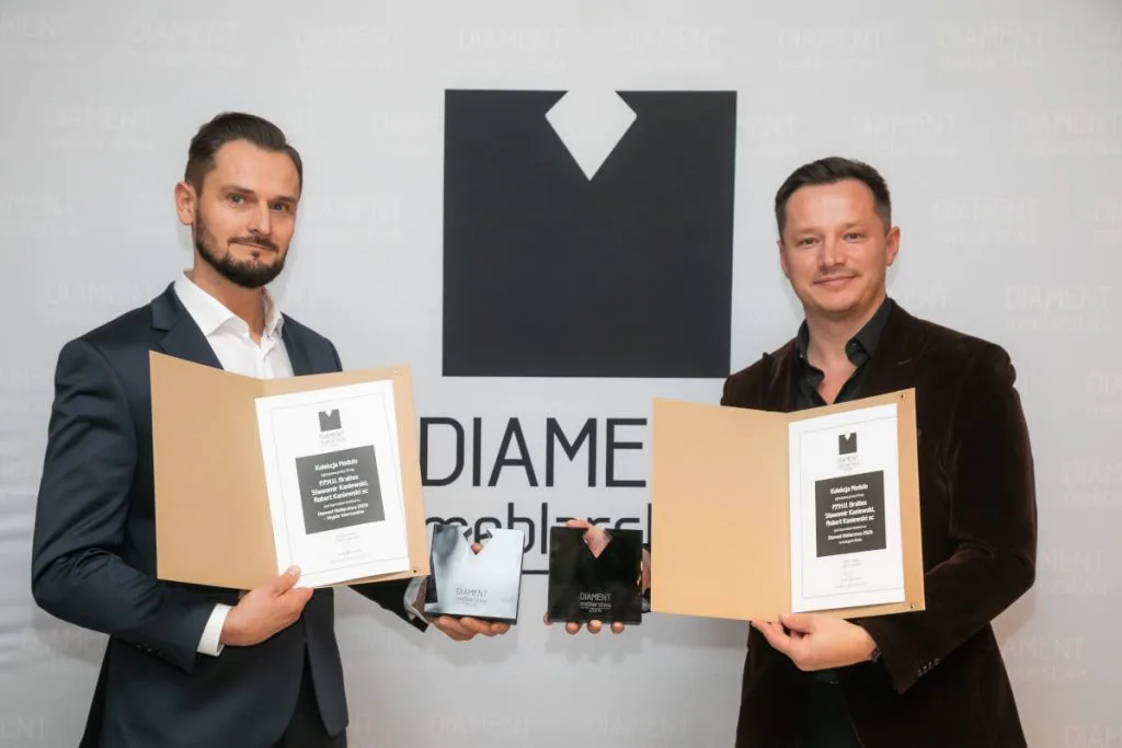 Dwaj mężczyźni prezentują otrzymane nagrody na tle ścianki z logo „DIAMENT meblarstwa”. Każdy z nich trzyma w dłoniach otwarty dyplom w teczce oraz czarną statuetkę. Napisy na dyplomach wskazują na nagrodę dla Kolekcji Modulo firmy Brattex.