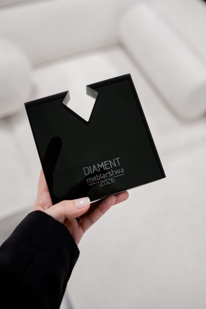 Zbliżenie na dłoń trzymającą czarną, akrylową statuetkę o kwadratowym kształcie z charakterystycznym wycięciem u góry. Na dole statuetki widnieje grawerowany napis „DIAMENT Meblarstwa 2026”.