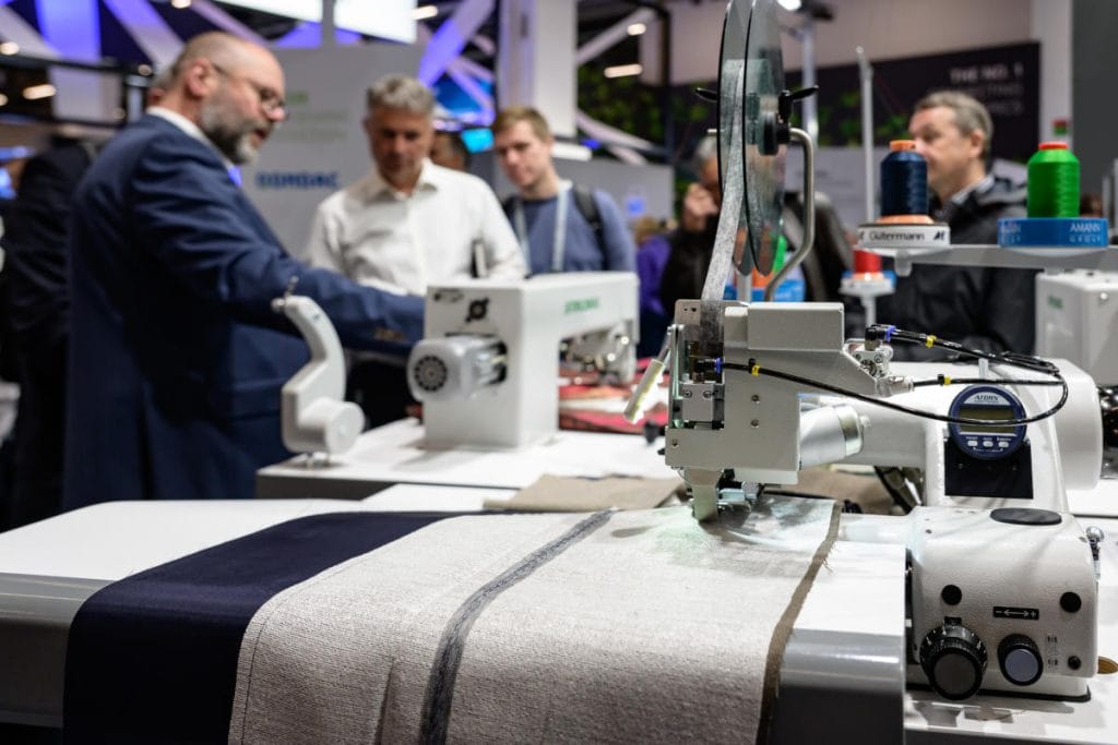 Zbliżenie na nowoczesną, przemysłową maszynę do szycia prezentowaną na stoisku targowym. Urządzenie pracuje nad jasnym materiałem z ciemniejszym pasem. W tle widać grupę mężczyzn w strojach biznesowych, którzy z zaciekawieniem obserwują pokaz technologii. Na maszynie widoczne są kolorowe szpulki nici: pomarańczowa i zielona.
